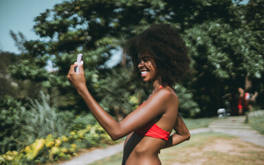 A laughing funny young African-American woman in a bikini is vlogging and making a broadcast via her smartphone or having a video call with a friend while standing on a beach area of a tropical resort