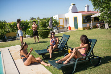 Content friends partying near swimming pool. Young women in swimsuits lying on deck chairs,...