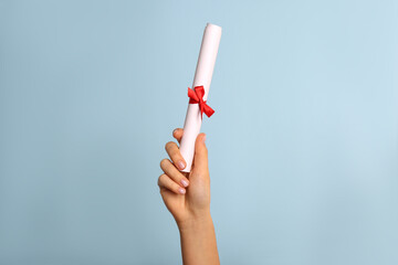 Student holding rolled diploma with red ribbon on light blue background, closeup - obrazy, fototapety, plakaty