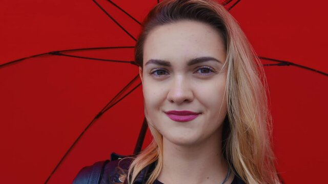 Beautiful girl under red umbrella looks at camera, blinks, smiles. Portrait of young caucasian woman, student. Closeup. Positive brown-eyed blonde lady. Front view. Gentle feminine glance
