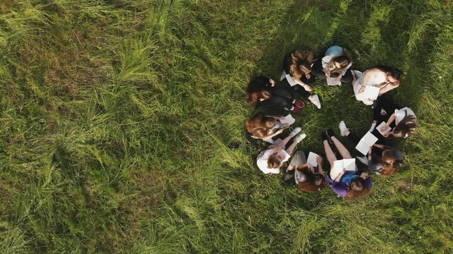 A group of 11 girls are sitting with notebooks on the grass in the park. Student work.