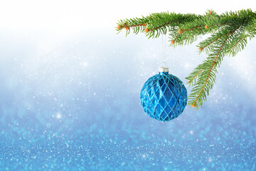 Christmas snowy decoration with hanging blue glitter ball and fir tree branch on blurred shiny blue bokeh background.