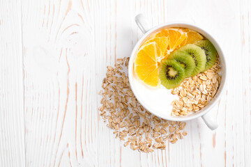 yogurt with kiwi and orange and oatmeal in a white bowl and with oatmeal grains on a white wooden background