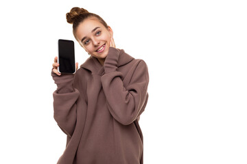 Photo of a beautiful happy cute girl with dark hair and a bun in a brown hoodie with a hood holding a mobile phone with a blank black screen isolated on a white background with free space for text