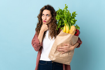 Young woman holding a grocery shopping bag isolated on blue background having doubts