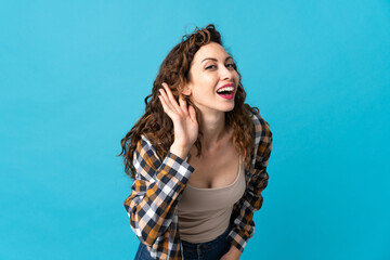 Young caucasian woman isolated on blue background listening to something by putting hand on the ear