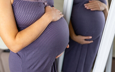 Expecting female wearing a purple nightgown and her reflection. Caring future mom holds her belly tenderly. The tummy on the last months of pregnancy close up.