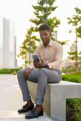 Fototapeta premium Portrait of young handsome African businessman sitting outdoors in city using mobile phone