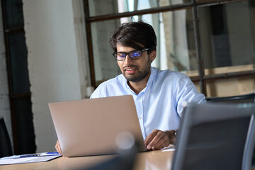 Young Indian businessman professional employee wearing eyeglasses using computer working online, e learning, watching business webinar in office, having virtual chat meeting looking at laptop at work.