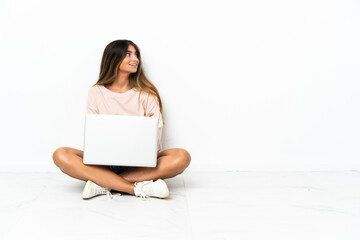 Young woman with a laptop sitting on the floor isolated on white background looking up while smiling
