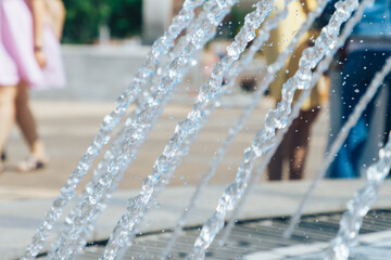 A splashing fountain in the center of the city with a selective focus and a blurry background of people walking. A running fountain with splashes and drops of water.