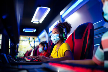 Portrait of an African American girl wearing face mask and headset playing video games on computer in game room during corona virus pandemic.
