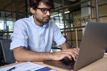 Young smart Indian business man employee wearing glasses using computer working online in office. Ethnic guy entrepreneur or manager typing on laptop analyzing digital data technology at workplace.