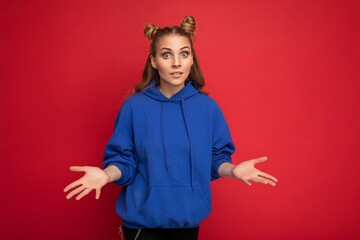 Photo of young charming beautiful attractive asking blonde woman with two horns with sincere emotions wearing stylish hipster blue hoodie isolated over red background with copy space and having