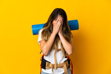 Young mountaineer woman with a big backpack isolated on yellow background with tired and sick expression