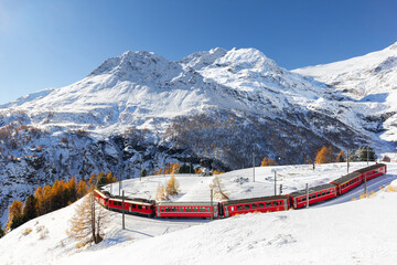 A red train is passing the train tracks with tight 180° curve at high Alp Grum. The Piz Palu peak...