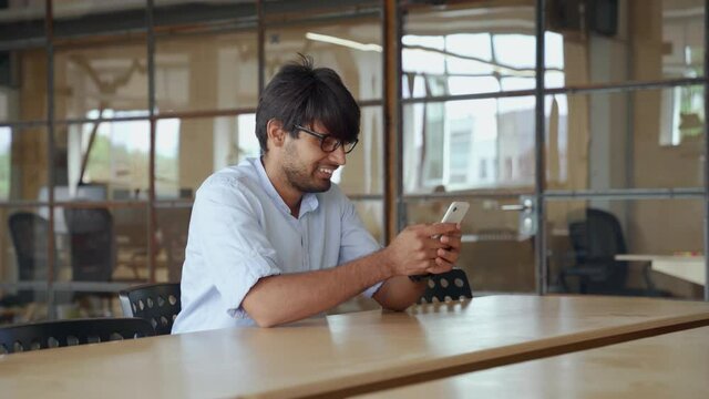 Smiling Indian business man using cellphone apps working in company office. Happy Arab professional businessman holding mobile phone using corporate digital data application technology concept.