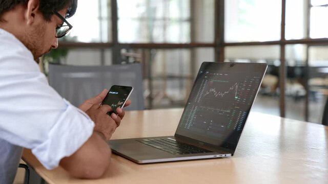 Indian business man trader broker investor analyst analyzing stock exchange market crypto trading chart data, managing financial risks looking at laptop using mobile app on smartphone.