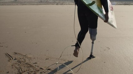 Surfer with disability holding surfboard under arm on ocean. Back view of man in diving suit walking along sandy beach carrying surfboard with foot tied to it with strap. Disability, sport concept