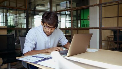 Young smart Indian business man employee wearing glasses using computer working online in office. Ethnic guy entrepreneur or manager typing on laptop analyzing digital data technology at workplace. - Powered by Adobe