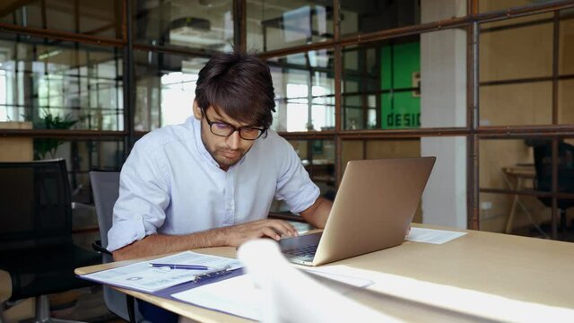 Young smart Indian business man employee wearing glasses using computer working online in office. Ethnic guy entrepreneur or manager typing on laptop analyzing digital data technology at workplace.