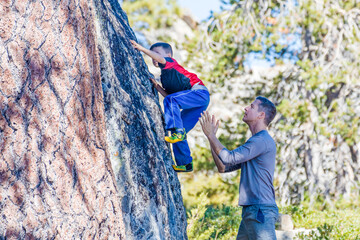 Young boy learning rock climbing techniques from his father who is close by and ready to catch his...