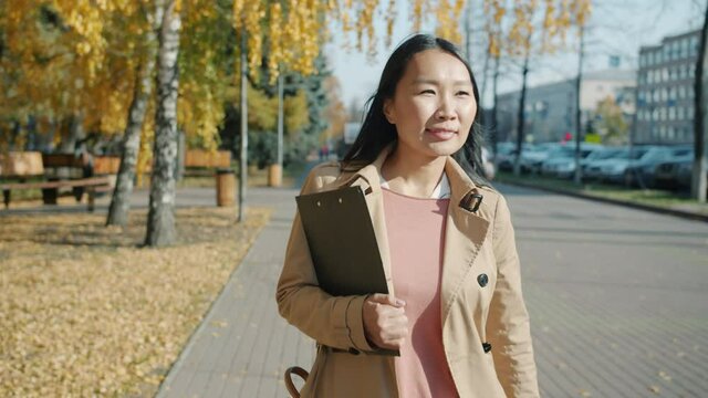 Dolly shot of young attractive Asian lady wearing elegant clothing walking outside in urban streen smiling carrying papers. People and city life concept.