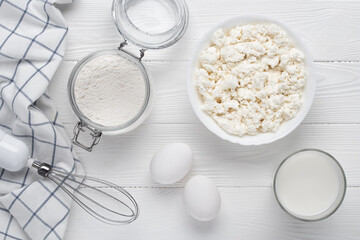 Raw curd cheese, flour, eggs, milk and whisk on a white wooden table. Cooking concept. Recipe ingredients. Baking products. View from above. Flat lay.