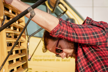 Young man in red plaid shirt and retro style with sunglasses leaning on the hood of a yellow bus