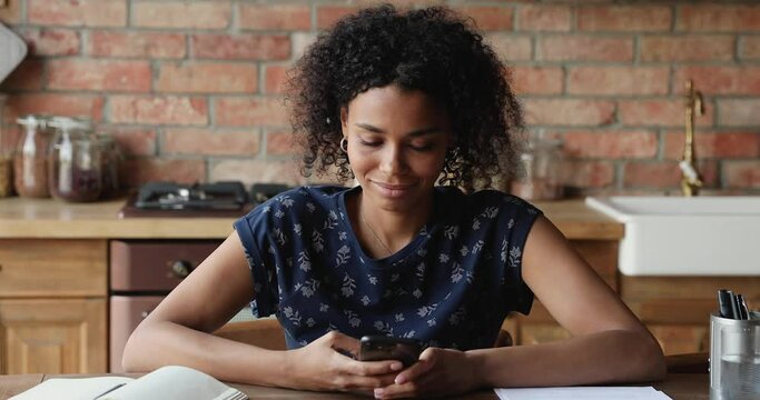 Attractive young African woman sit at table in kitchen holds smartphone texting browsing social media, share messages through messenger, spend free time on internet, buying, chatting remotely concept