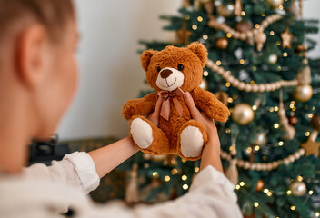 Merry Christmas and Happy New Year! A girl holding a toy plush teddy bear on the background of a decorated Christmas tree.