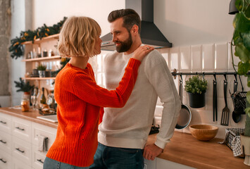 Merry Christmas and Happy New Year! A loving cute young couple standing in the kitchen and embracing, which is decorated with branches of a Christmas tree for the upcoming holiday.