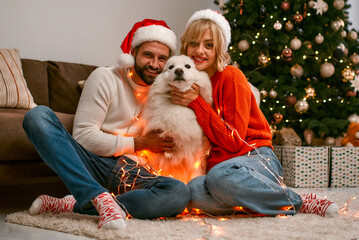 Merry Christmas and Happy New Year! Happy couple with dog  is waiting for the New Year together in Santa Claus hats while sitting near beautiful Christmas tree at home.