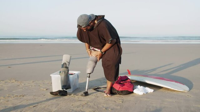 Surfer with disability putting on prosthesis on coastline. Long shot of bearded Caucasian man in cap making preparation for surf training in ocean on summer morning. Disability, surfing concept