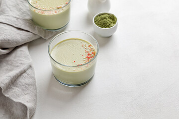 Two matcha panna cotta desserts with confectionery decor in a glass on a white background and a grey napkin next to it.