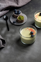 Matcha panna cotta dessert with confectionery red decor with blueberry in a glass with spoon on grey background. Matcha tea next to it. Minimalistic photography.