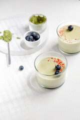 Matcha panna cotta dessert with confectionery red decor with blueberry in a glass with spoon on white background. Matcha tea next to it. Minimalistic and sunny photography.