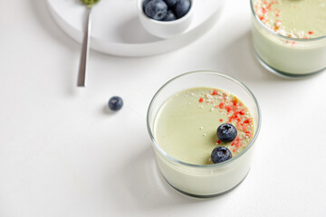 Matcha panna cotta dessert with confectionery red decor with blueberry in a glass with spoon on white background. Matcha tea next to it. Minimalistic and sunny photography.