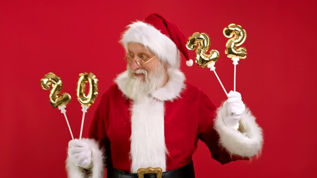 Merry Santa Claus is Enjoying Coming Christmas Holding Inflatable Gold Numbers 2022 in His Hands, Dancing and Having Fun on Red Background. Happy New Year 2022. Merry Christmas and Happy New Year.