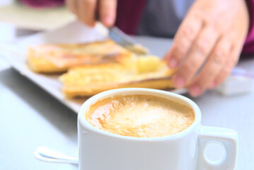 Espresso macchiato Coffee cup. Bread and butter toast out of focus at the background with hands and knife