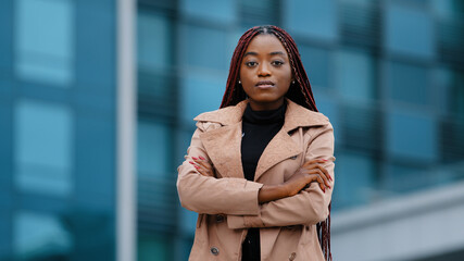 Serious concentrated young african american girl with afro hairstyle standing with folded hands, portrait outdoor. Focused black female professional looking at camera, on office building background