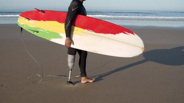 Man with artificial leg walking along beach with surfboard. Side view of surfer with disability in diving suit going to ocean for training, holding surfboard under arm. Disability, water sport concept