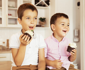 Funny two little brothers are sitting together at the kitchen table and eating cupcakes.