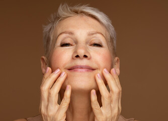 Closeup face of mature woman with wrinkles massaging face isolated over brown background. Aging...