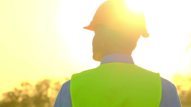Engineer in uniform on windmill station. Skilled worker in orange hardhat walks towards rotating wind turbine illuminated by shining sun backside view