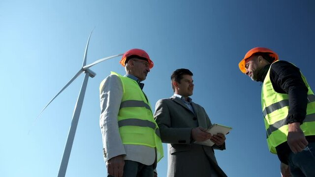 Wind driven generator produces energy against blue sky. Manager and engineers in helmets check project documentation on tablet low angle shot