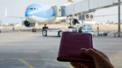 A woman holds a passport against the background of an airplane at the airport. The faceless girl is waiting for her flight and looks at the airfield through the window.
