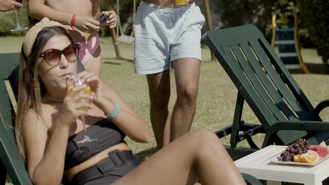 Carefree young people in swimwear relaxing in backyard. Attractive Caucasian men and women spending weekends together, drinking cocktails, eating fruits, sunbathing. Summer concept