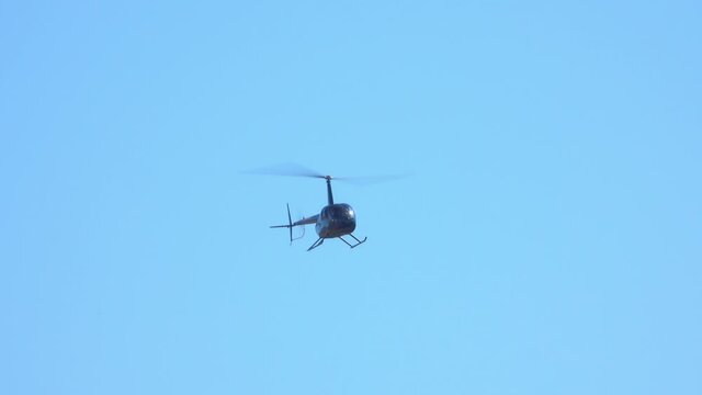 A small, black helicopter is shown slowly approaching, set against a clear blue sky.
