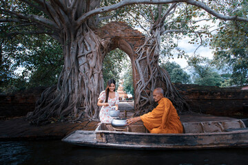  Young woman give alms to a Buddhist monk at front of ancient gate of Pra Ngam temple under Bodhi...